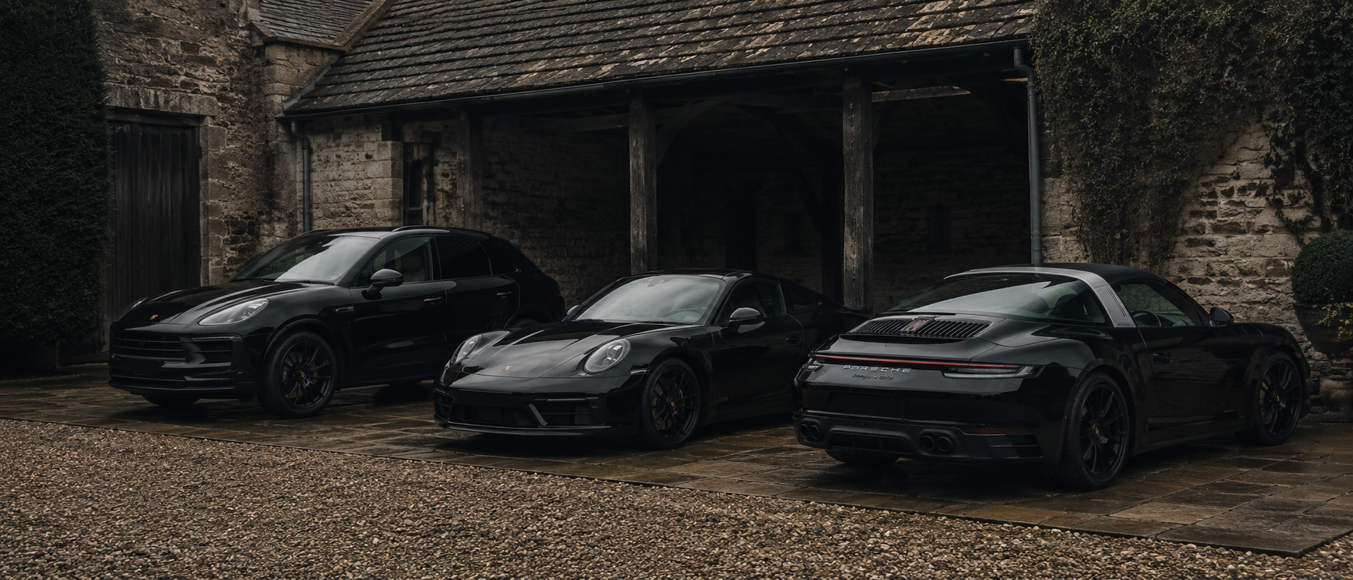 The three cars arranged on a stone-flagged forecourt in front of a timber-framed coach house: Macan GTS on the left, 911 Carrera GTS in the centre, 911 Targa 4 GTS on the right.