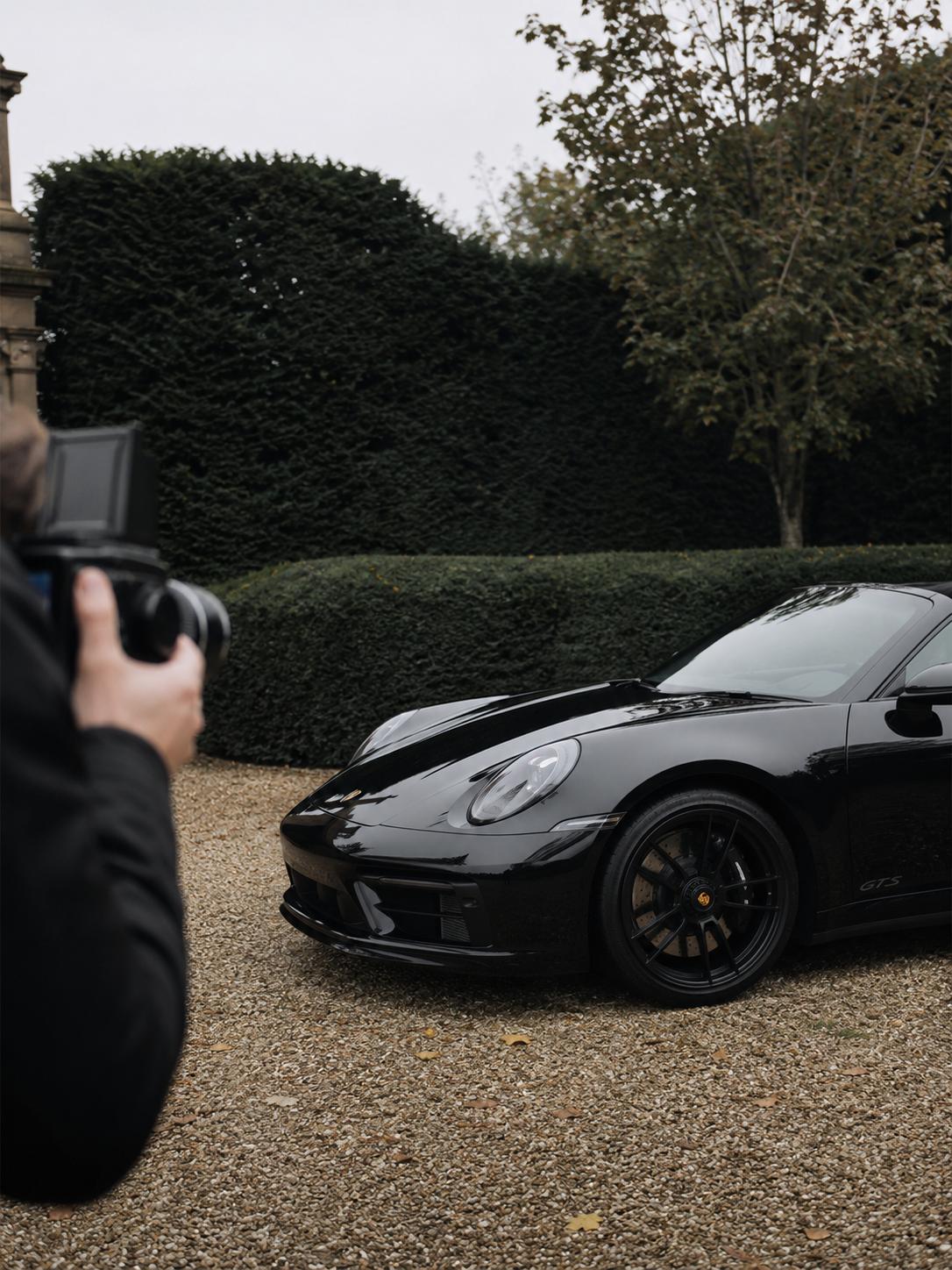 A photographer in the foreground frames a black Porsche 911 parked on a gravel forecourt in front of dark green hedging.